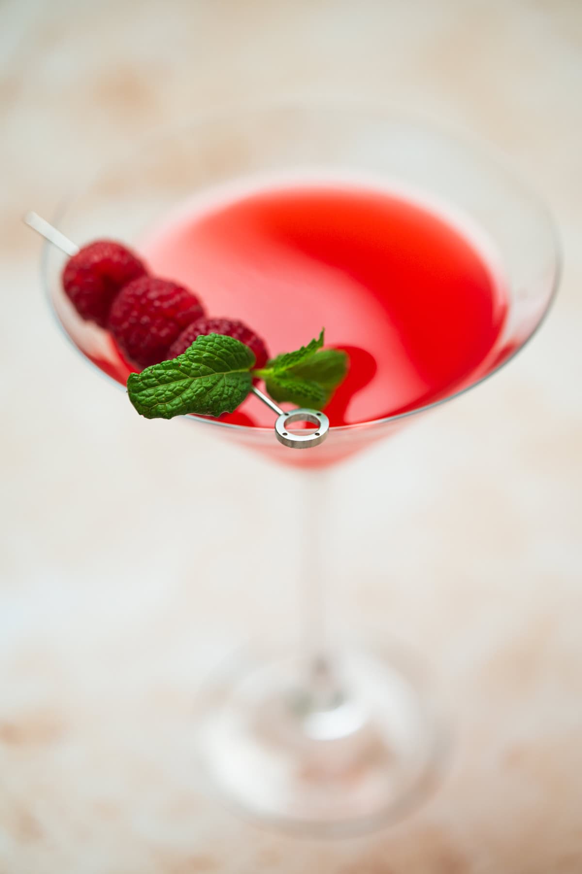 A vibrant raspberry mint martini in a martini glass, garnished with fresh raspberries and a sprig of mint on a cocktail pick, set against a soft, blurred background.