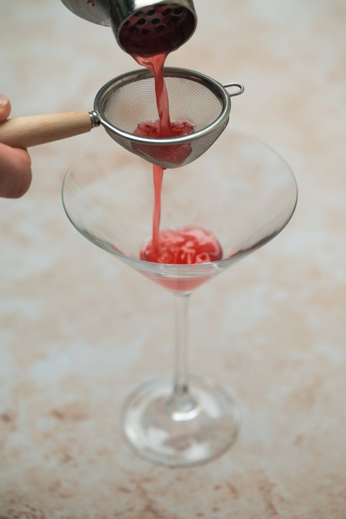 Red liquid for a raspberry mint martini is poured through a fine mesh strainer into a clear, empty martini glass on a light, textured surface.