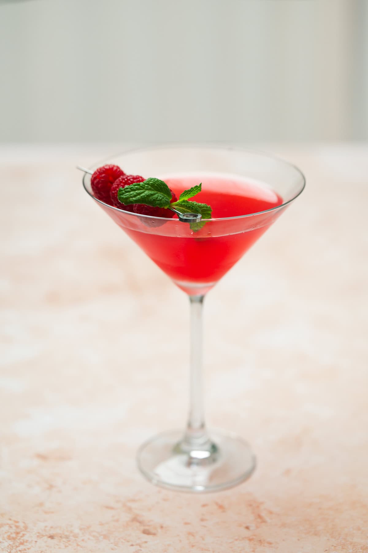 A raspberry mint martini, served in a martini glass and garnished with fresh raspberries and a sprig of mint, sits on a light, textured surface.