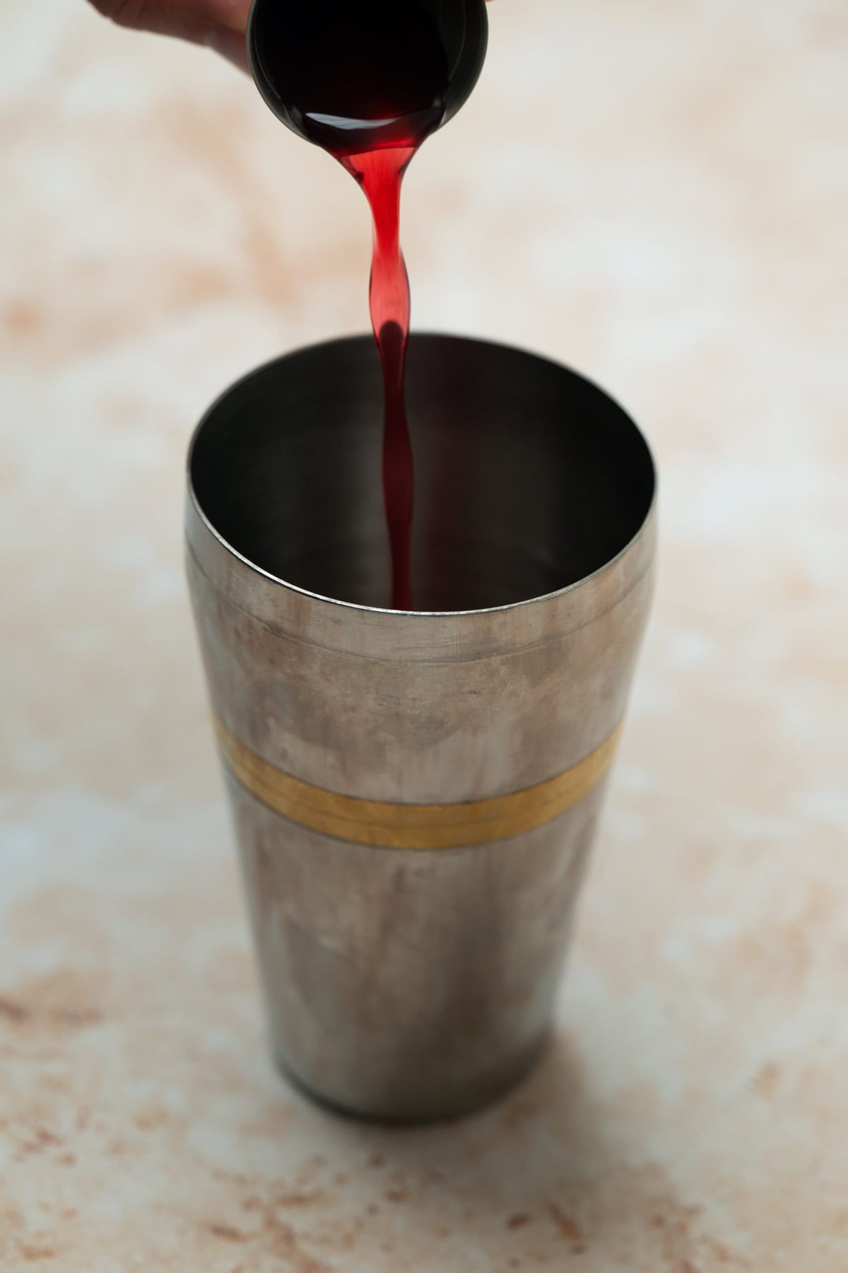 A hand pours red liquid for a raspberry mint martini from a small black cup into a stainless steel cocktail shaker with a gold band, set on a light-colored surface.