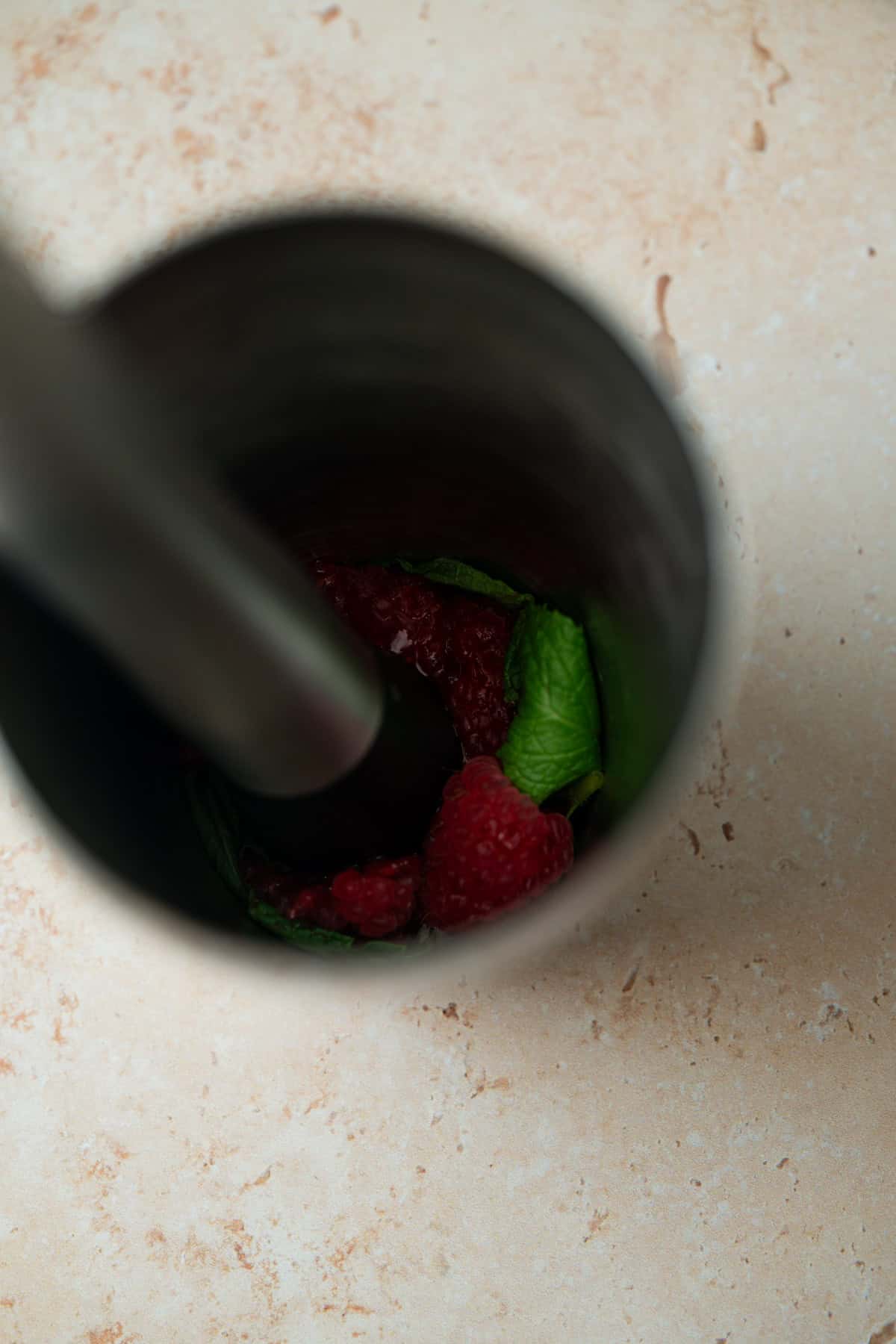 A metal muddler pressing fresh raspberries and mint leaves inside a metal cup, viewed from above on a light-colored countertop—perfect for crafting a refreshing raspberry mint martini.