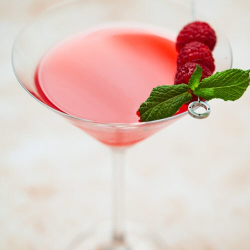 A martini glass filled with a bright pink raspberry mint martini, garnished with three raspberries and a sprig of fresh mint on a cocktail pick, set against a light, blurred background.