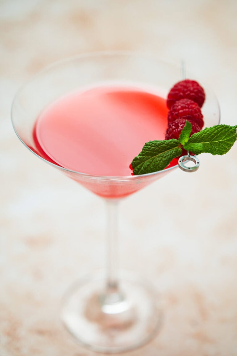 A martini glass filled with a bright pink raspberry mint martini, garnished with three raspberries and a sprig of fresh mint on a cocktail pick, set against a light, blurred background.