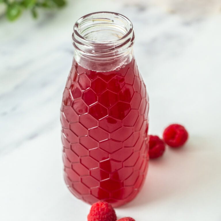 A glass bottle with a honeycomb pattern contains raspberry simple syrup. Three fresh raspberries are placed beside the bottle on a white surface, with the background softly blurred.