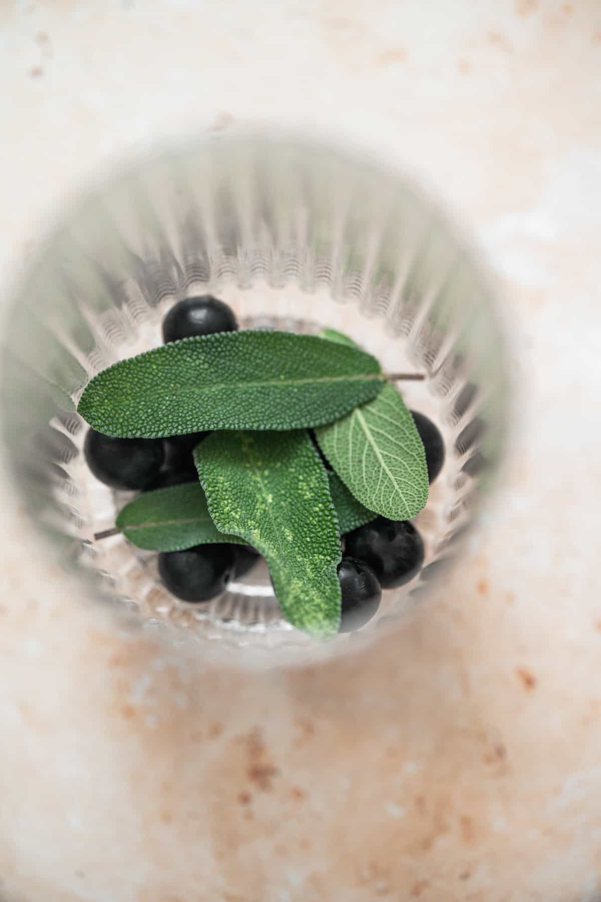 A glass viewed from above contains dark berries and a few fresh, textured green sage leaves, placed on a light beige stone surface—an inviting Blueberry Sage Cocktail ready to enjoy.