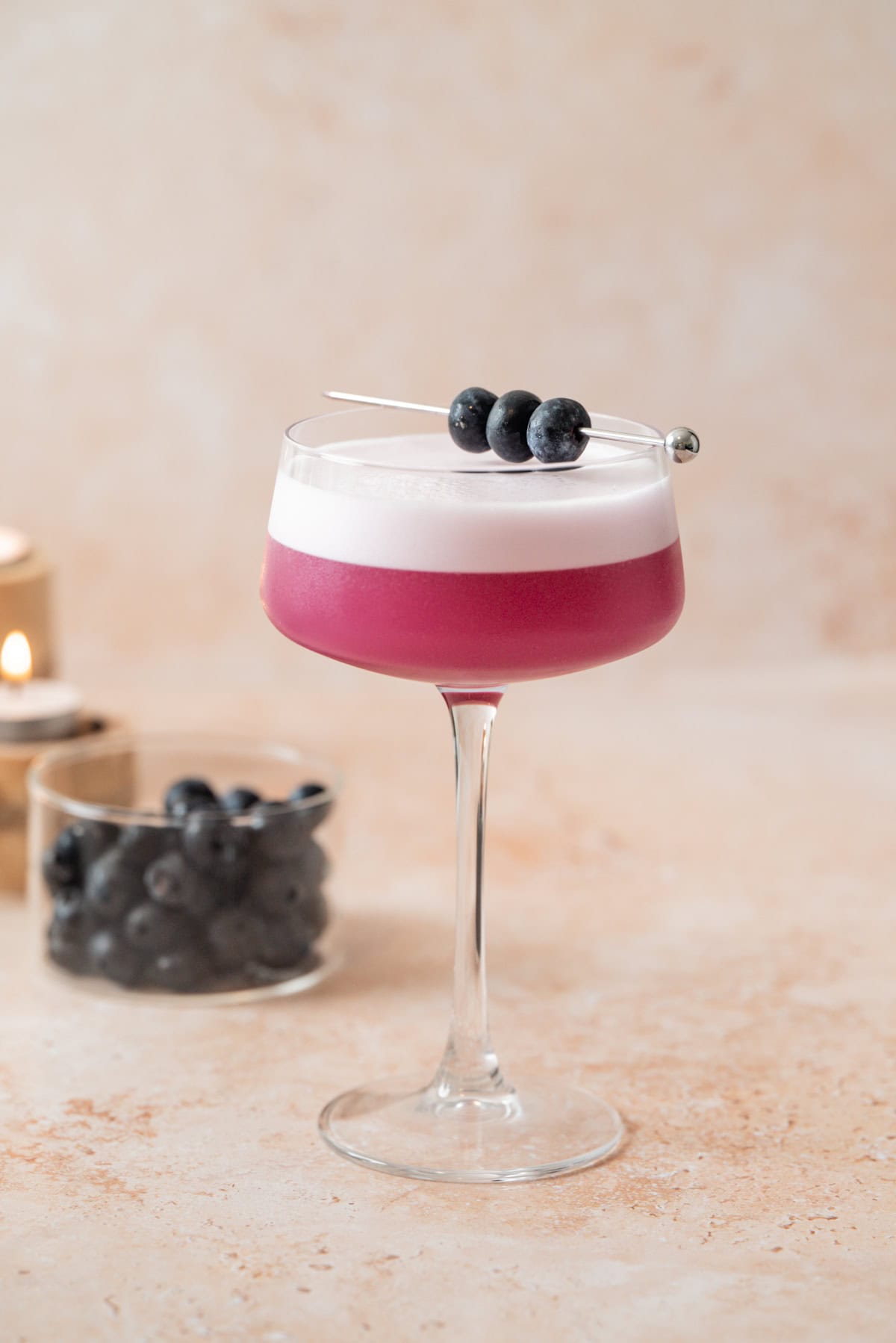 A purple Blueberry Gin Sour with a foamy top served in a coupe glass, garnished with three blueberries on a cocktail pick. A small glass bowl of blueberries and a lit candle are in the background.