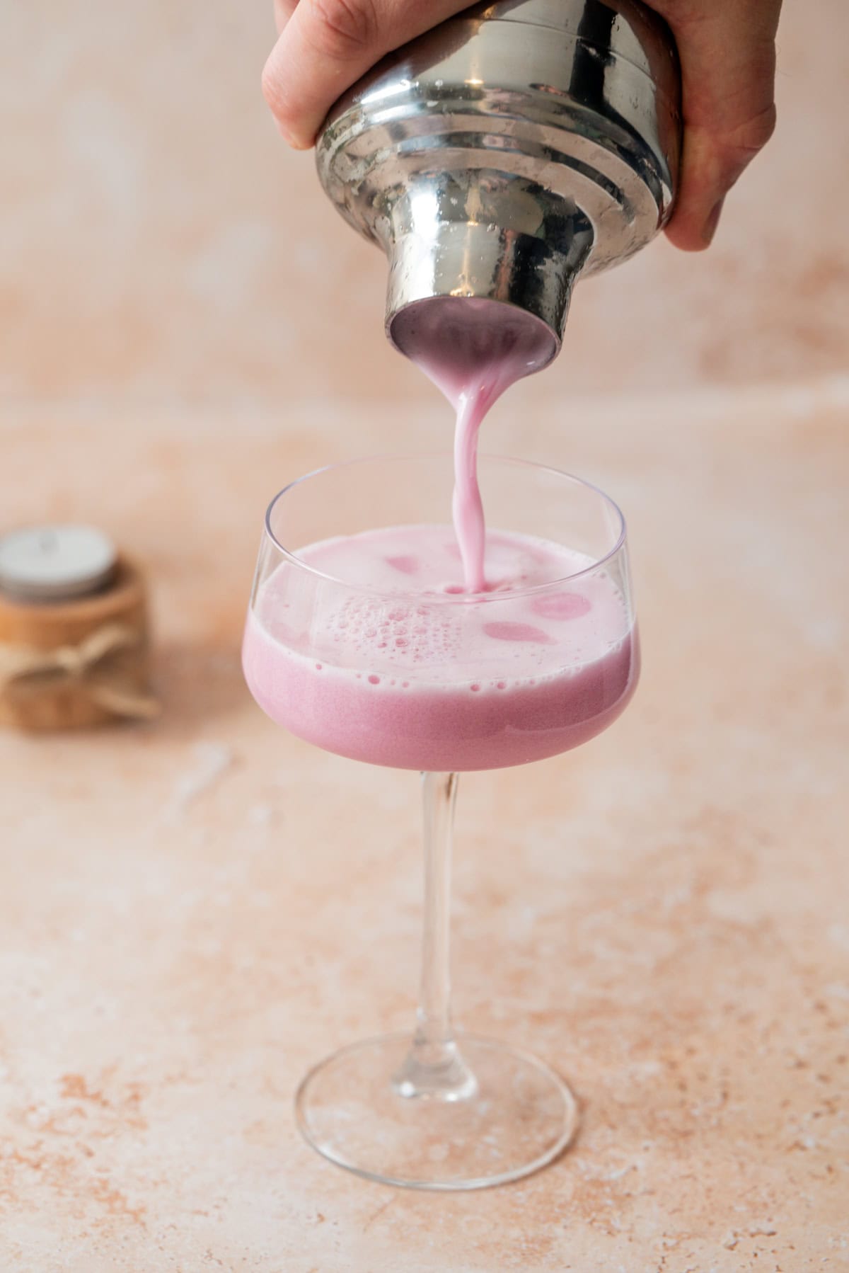 A hand pours a creamy, pink-purple Blueberry Gin Sour from a metal shaker into a stemmed glass, set on a light beige surface with a small jar in the background.