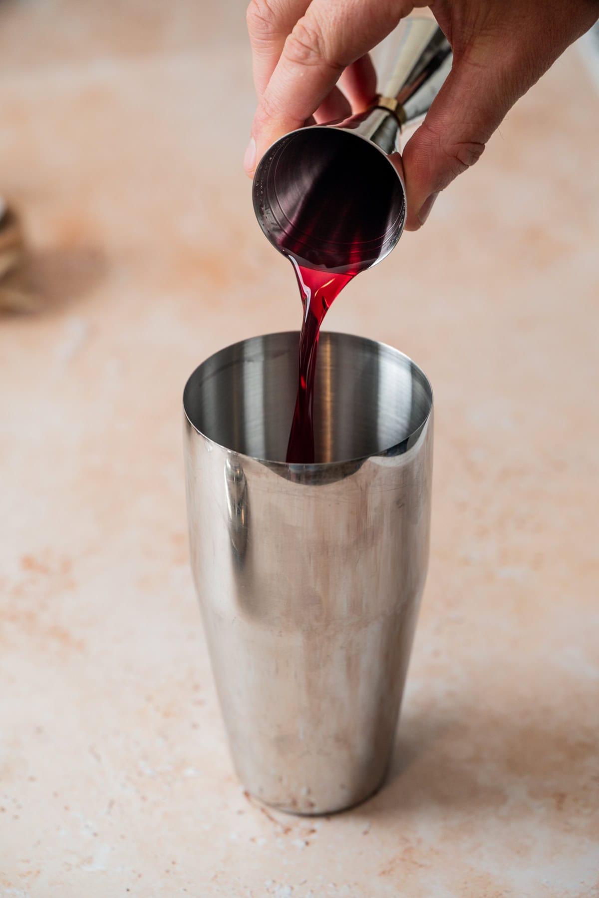 A hand pours red liquid for a Blueberry Gin Sour from a jigger into a stainless steel cocktail shaker on a light-colored surface.