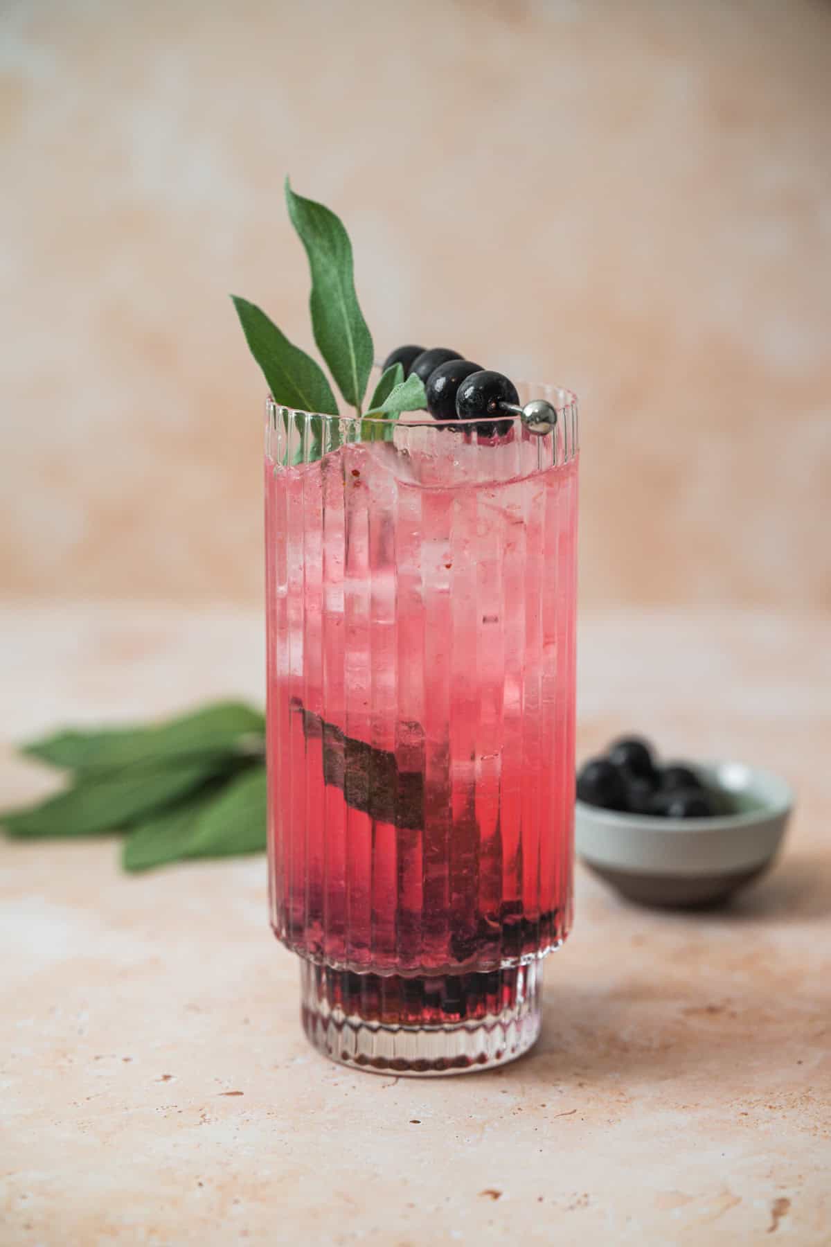 A tall, ribbed glass filled with a pink iced Blueberry Sage Cocktail, garnished with green leaves and dark berries on a skewer. A small bowl of berries and fresh leaves are placed in the background on a light, textured surface.