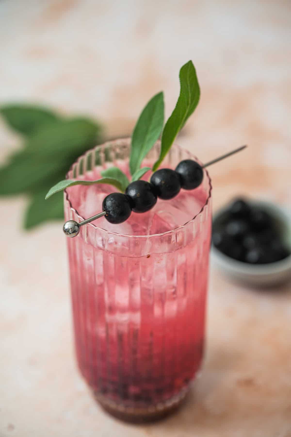 A tall glass of Blueberry Sage Cocktail over ice, garnished with three blueberries and a green leaf on a metal pick. A small bowl of blueberries and extra sage leaves are blurred in the background.