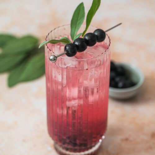 A tall, ribbed glass filled with a pink iced Blueberry Sage Cocktail, garnished with three dark berries and a sprig of green leaves on a cocktail pick. A small bowl of berries and sage leaves are in the background.