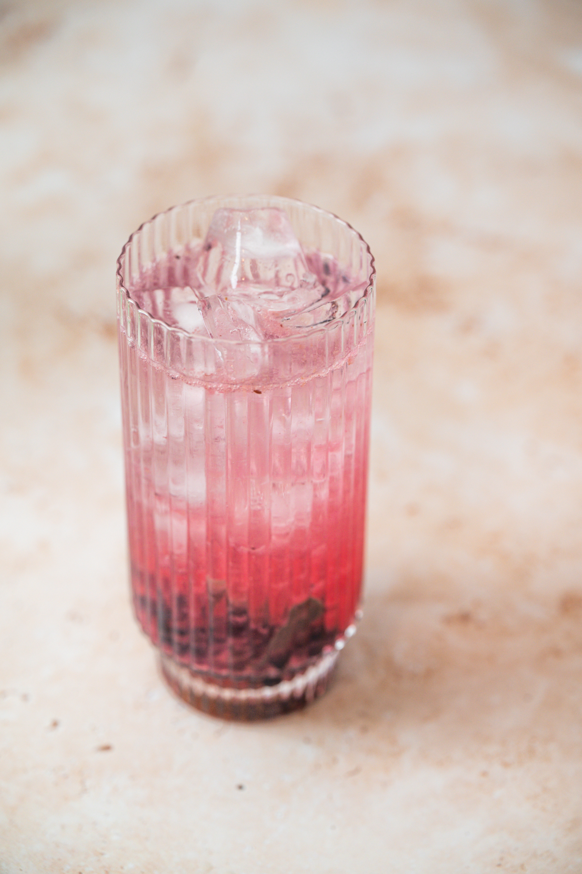 A tall, ribbed glass filled with a vibrant Blueberry Sage Cocktail, boasting pink and purple hues and visible crushed berries at the bottom, sits on a light marble surface.