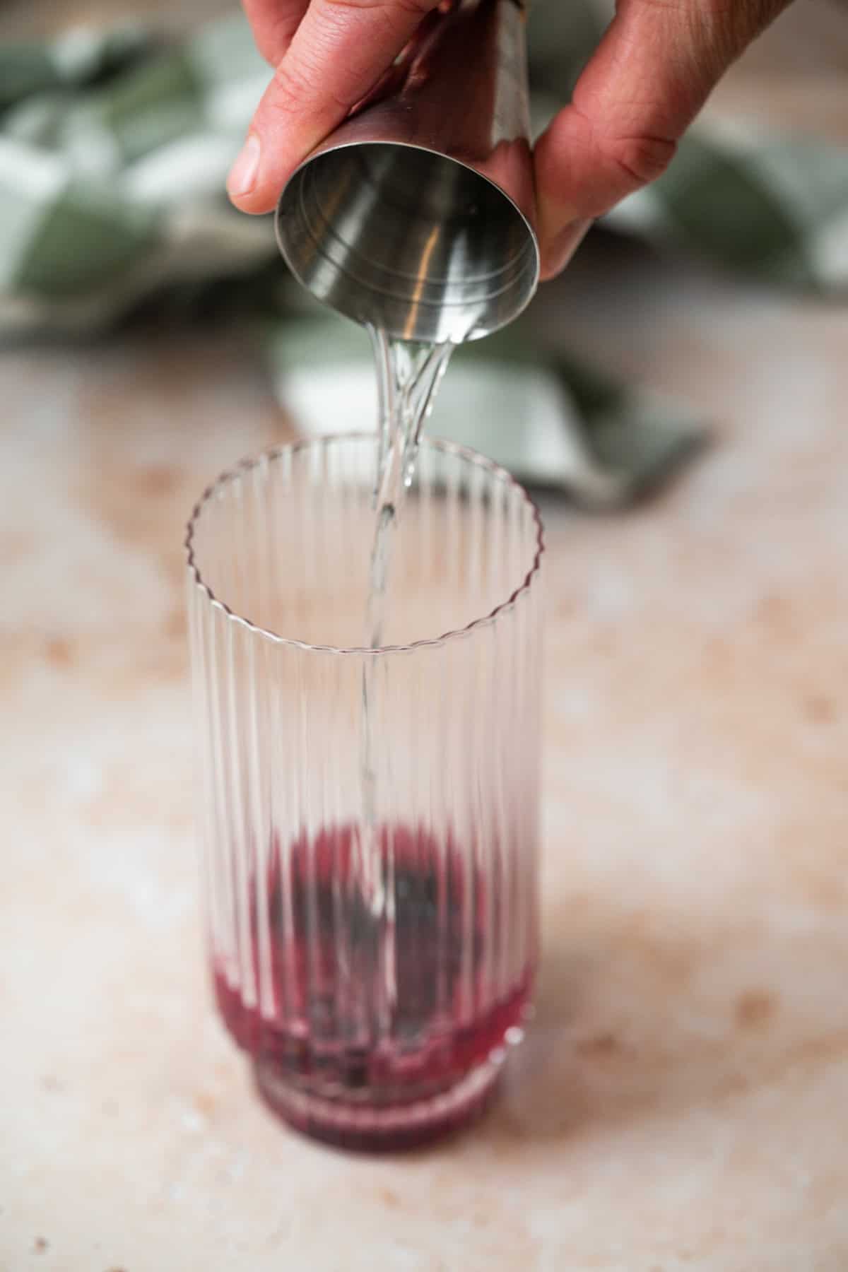 A hand pours clear liquid from a metal jigger into a tall glass containing a small amount of red Blueberry Sage Cocktail, set on a light-colored surface with a blurred cloth in the background.