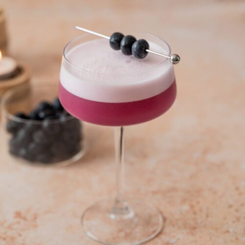 A coupe glass filled with a vibrant Blueberry Gin Sour cocktail, topped with a layer of foam and garnished with three blueberries on a cocktail pick. In the background, a small glass jar of blueberries sits on a light, textured surface.