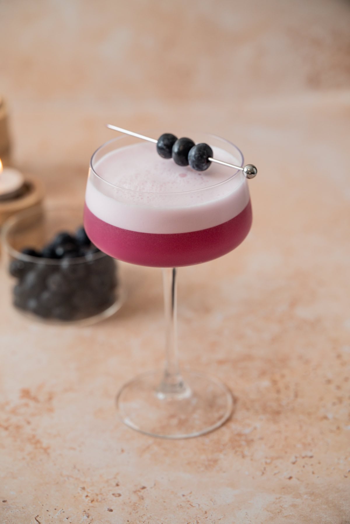 A coupe glass filled with a vibrant Blueberry Gin Sour cocktail, topped with a layer of foam and garnished with three blueberries on a cocktail pick. In the background, a small glass jar of blueberries sits on a light, textured surface.