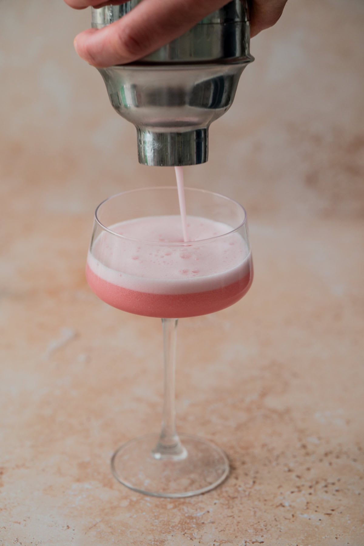 A hand pours a frothy raspberry gin sour from a metal shaker into a stemmed glass on a light, textured surface.