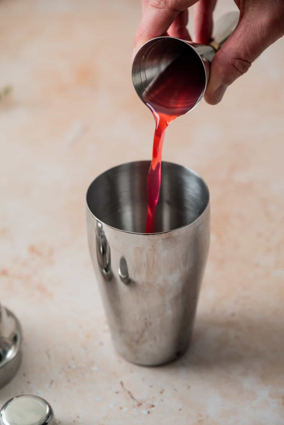 A hand pours a red liquid for a raspberry gin sour from a small metal measuring cup into a stainless steel cocktail shaker on a light-colored surface.