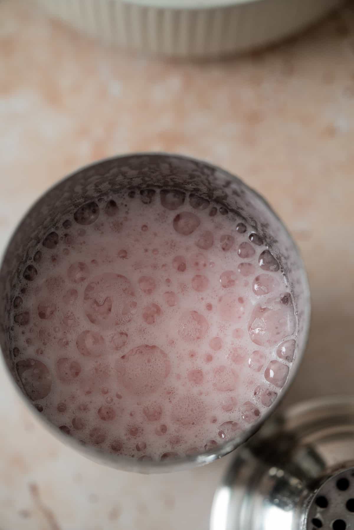 A close-up view of a metal cocktail shaker filled with a frothy, pink raspberry gin sour covered in bubbles, sitting on a beige surface.