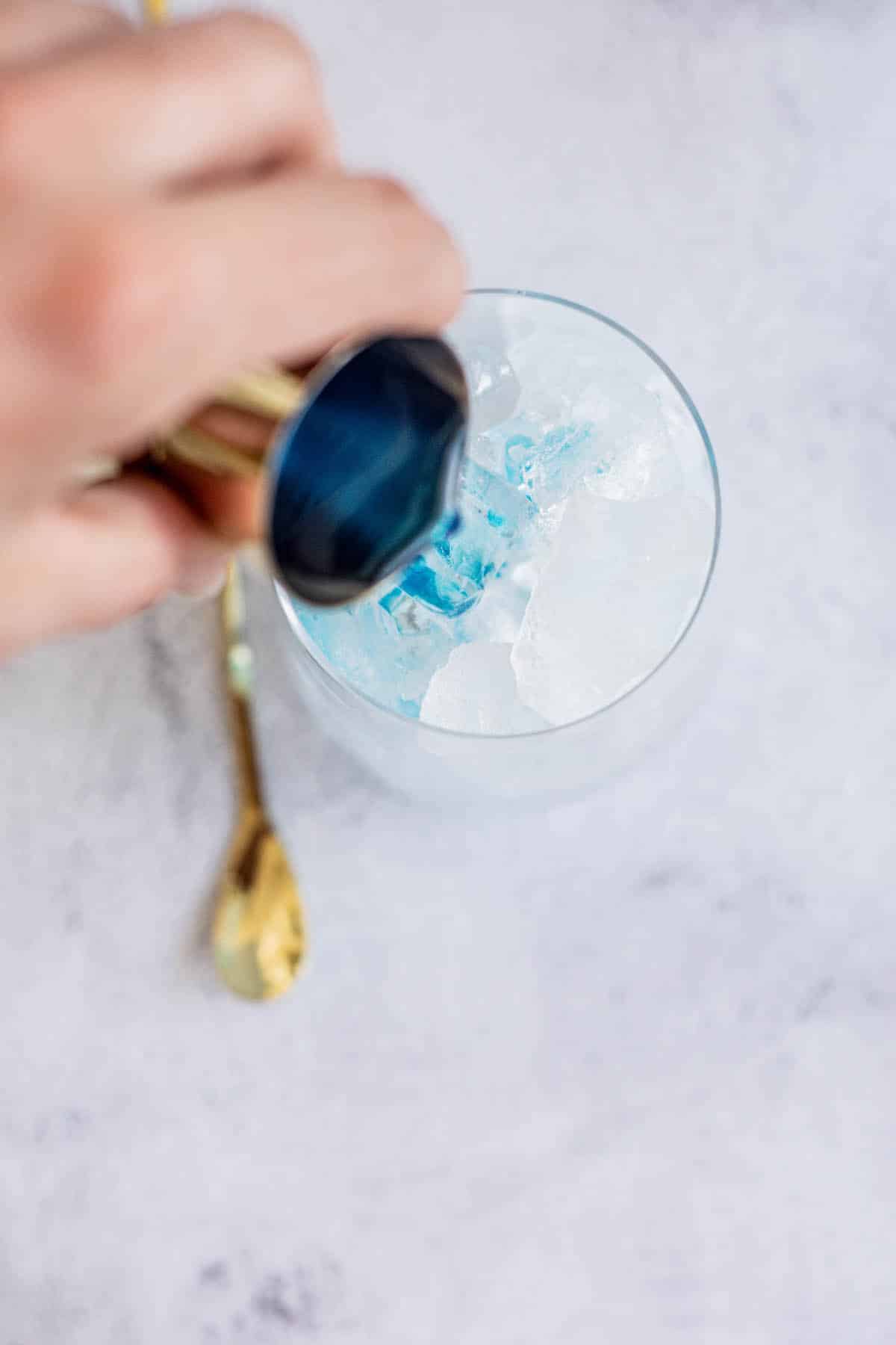 blue curacao syrup being in a tumbler being poured into a tall glass filled with ice next to a gold cocktail stirrer.