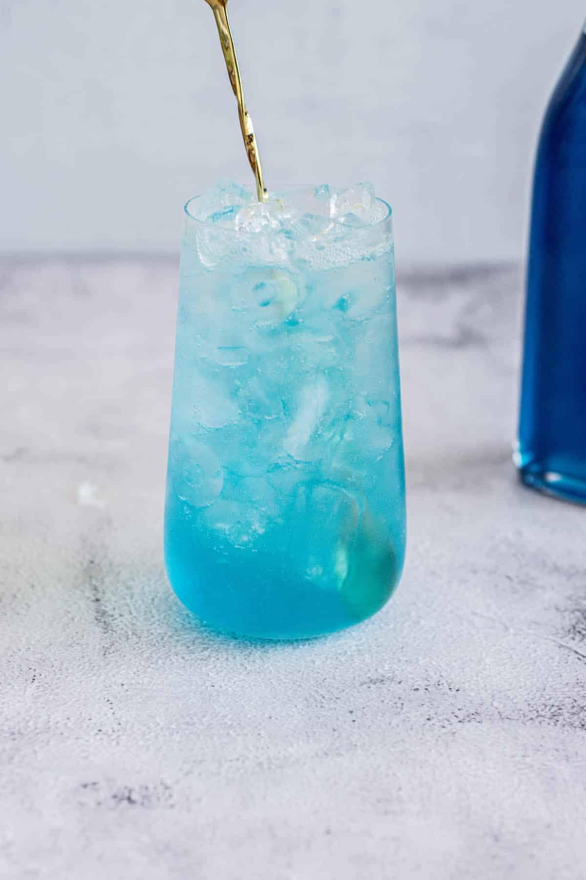 a blue lagoon mocktail in a tall glass with golden cocktail stirrer, with a bottle of blue Curaçao syrup behind.