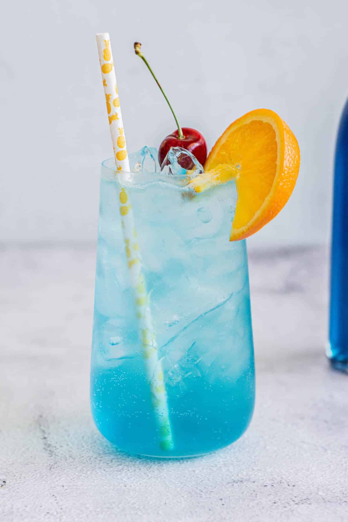 a blue lagoon mocktail in a tall glass with cherry, orange slice on rim and a straw, with a bottle of blue Curaçao syrup behind.