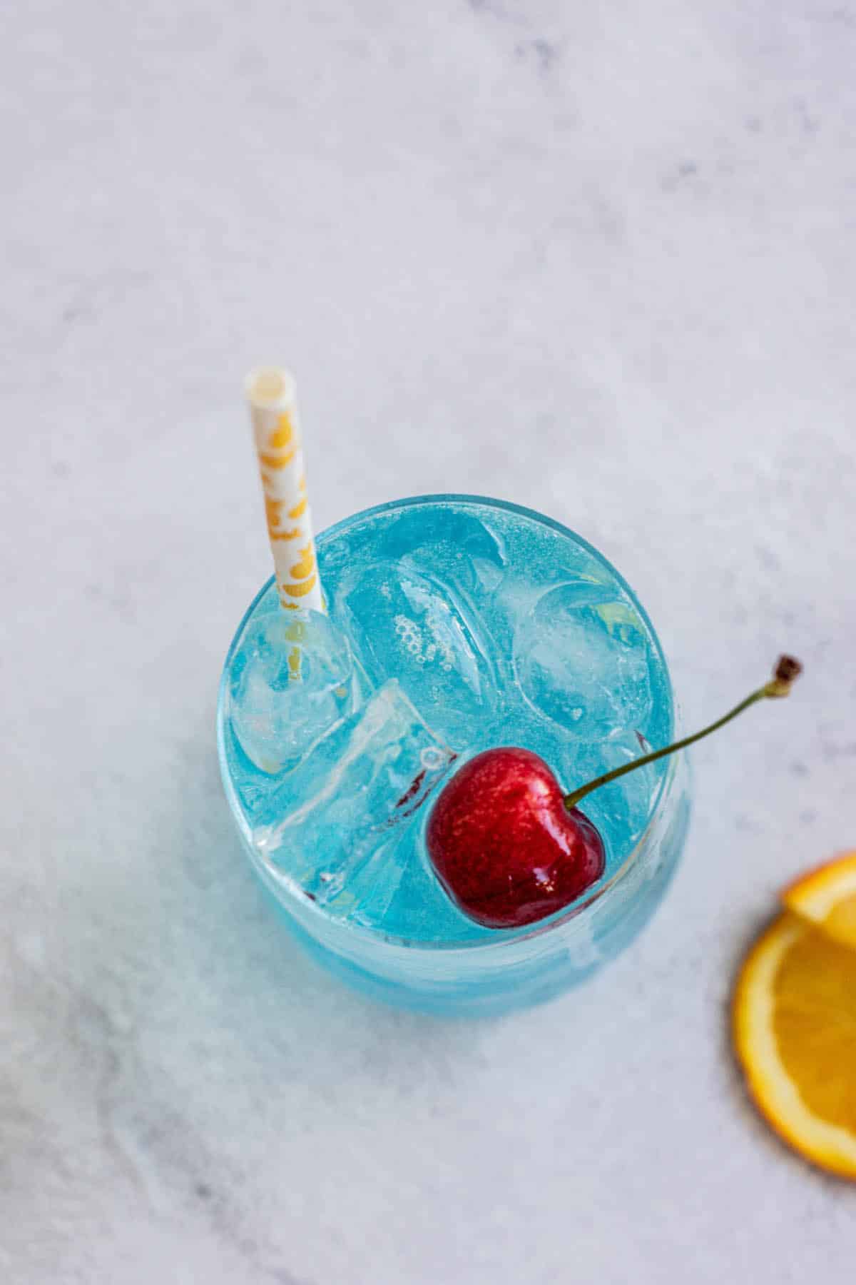 Birdseye view of a blue lagoon mocktail in a tall glass with cherry, orange slice next to glass and a straw.