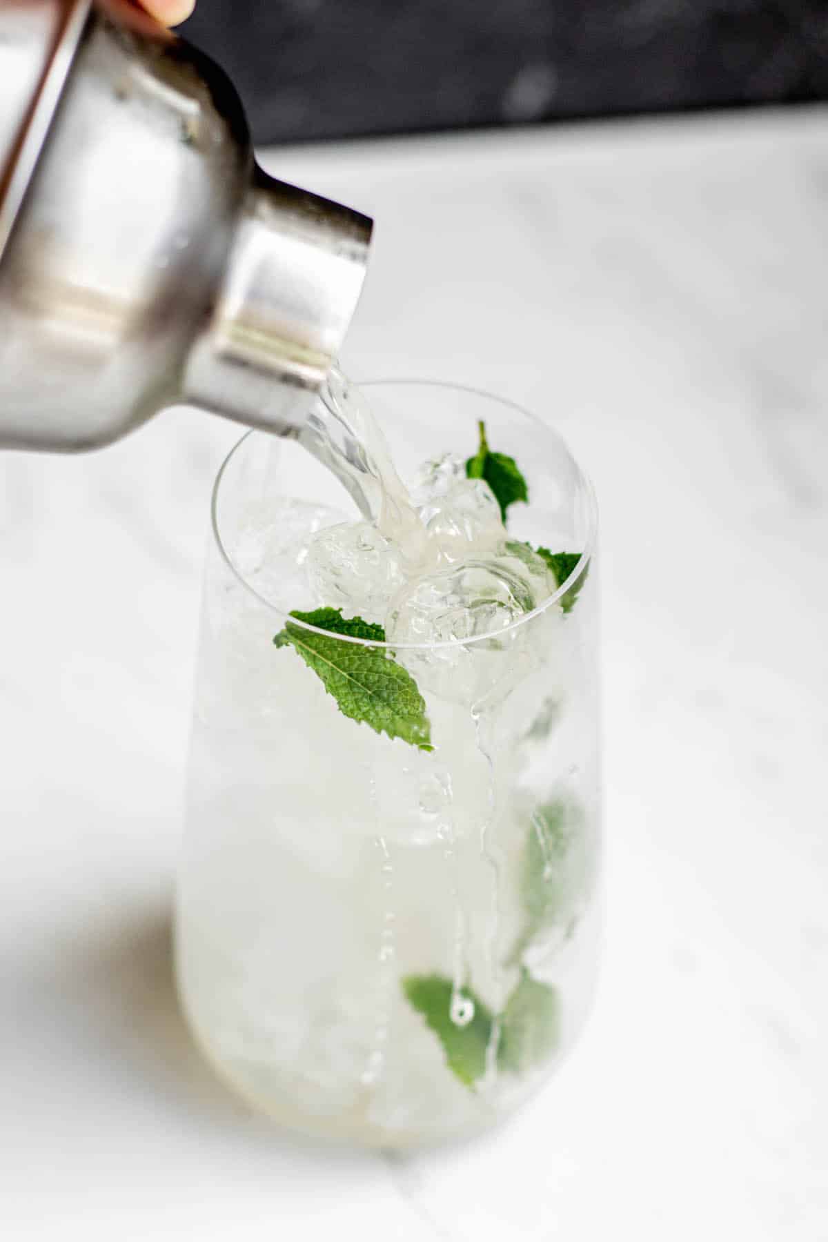 lychee mojito being poured into a tall glass with mint and ice from a cocktail shaker.