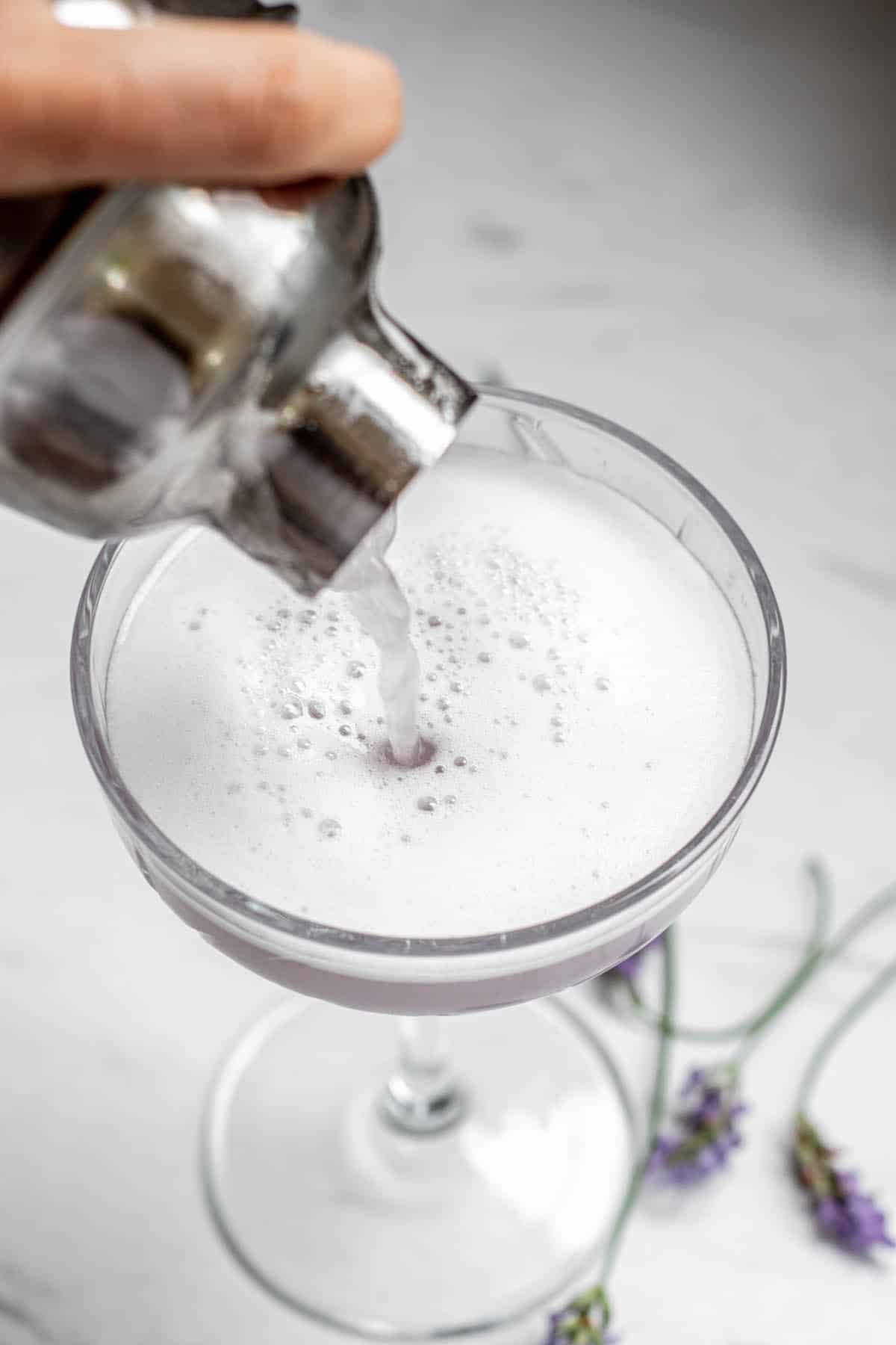 lavender gin sour being poured from a cocktail shaker and into a martini glass.