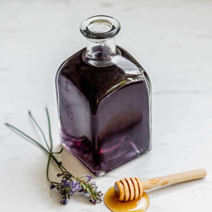 small glass bottle of honey lavender syrup with lavender and honey utensil next to it.