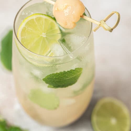 A glass of Lychee Mojito, iced and garnished with lime slices, fresh mint leaves, and a skewered lychee on top. A halved lime and mint leaves rest on the light-colored surface beside the glass.