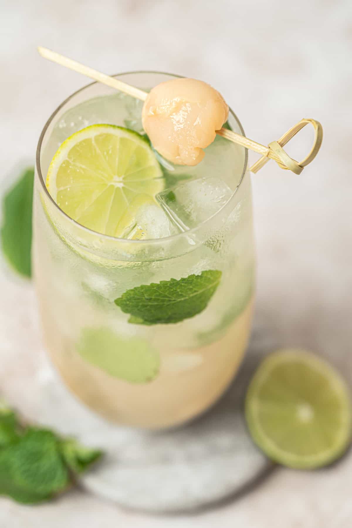 A glass of Lychee Mojito, iced and garnished with lime slices, fresh mint leaves, and a skewered lychee on top. A halved lime and mint leaves rest on the light-colored surface beside the glass.