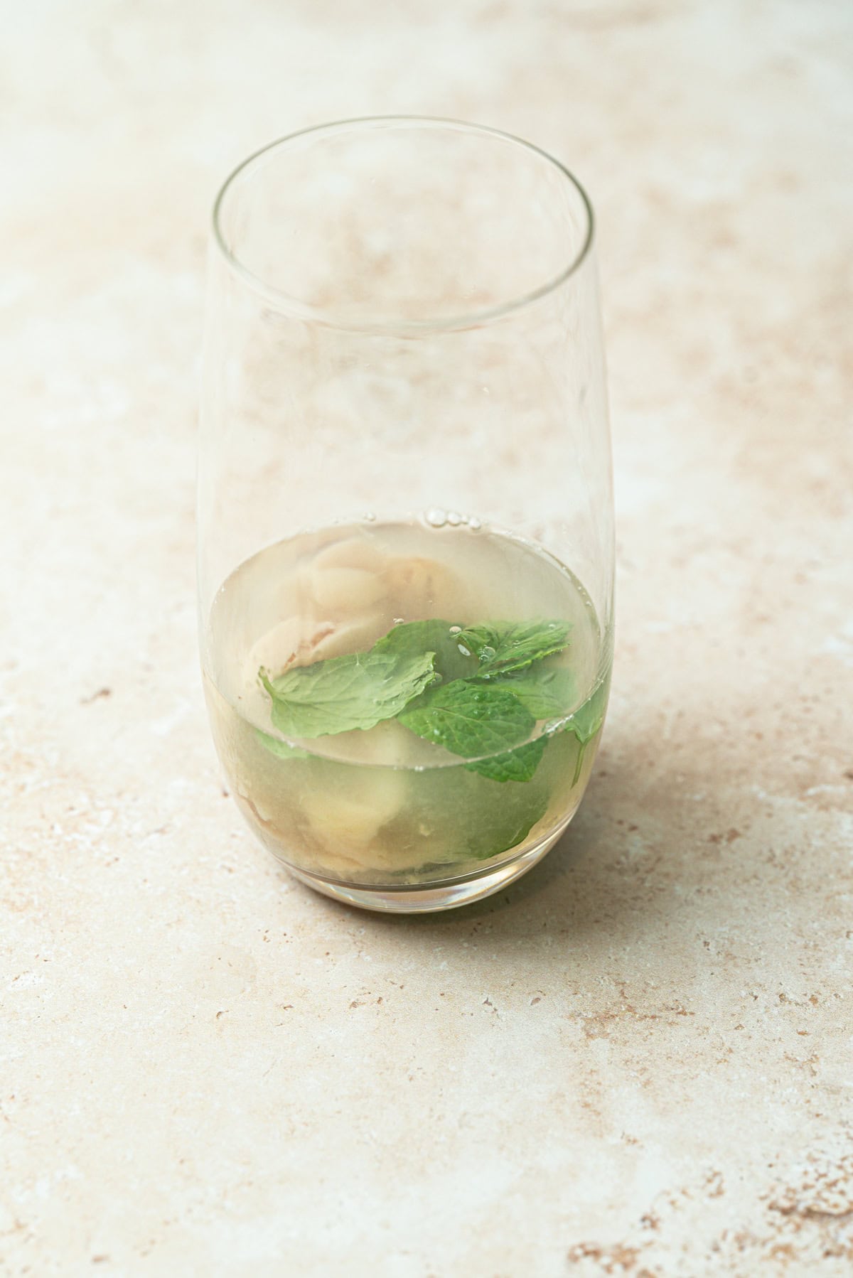 A clear glass of Lychee Mojito with a light, slightly cloudy drink, lychee fruits, and fresh mint leaves sits on a beige textured surface.