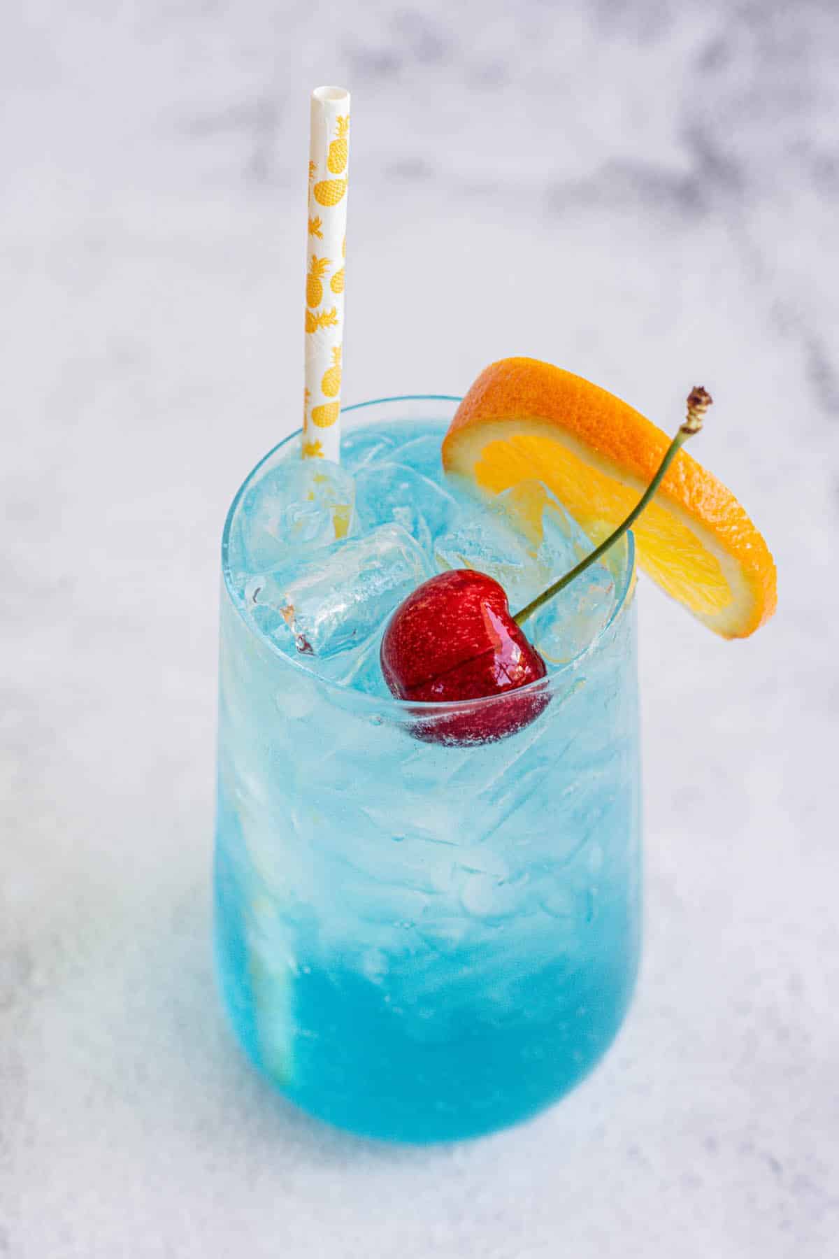 top down view of a blue lagoon mocktail in a tall glass with cherry, orange slice on rim and a straw.