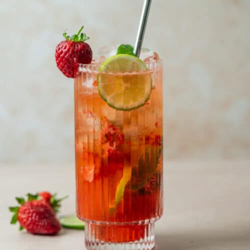 A tall, ribbed glass filled with a vibrant Strawberry Mojito, garnished with a lime slice, a whole strawberry, and a metal straw. Fresh strawberries and a lime wedge are placed beside the glass on a light surface.