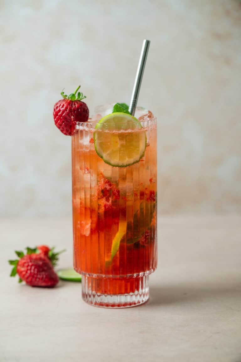 A tall, ribbed glass filled with a vibrant Strawberry Mojito, garnished with a lime slice, a whole strawberry, and a metal straw. Fresh strawberries and a lime wedge are placed beside the glass on a light surface.