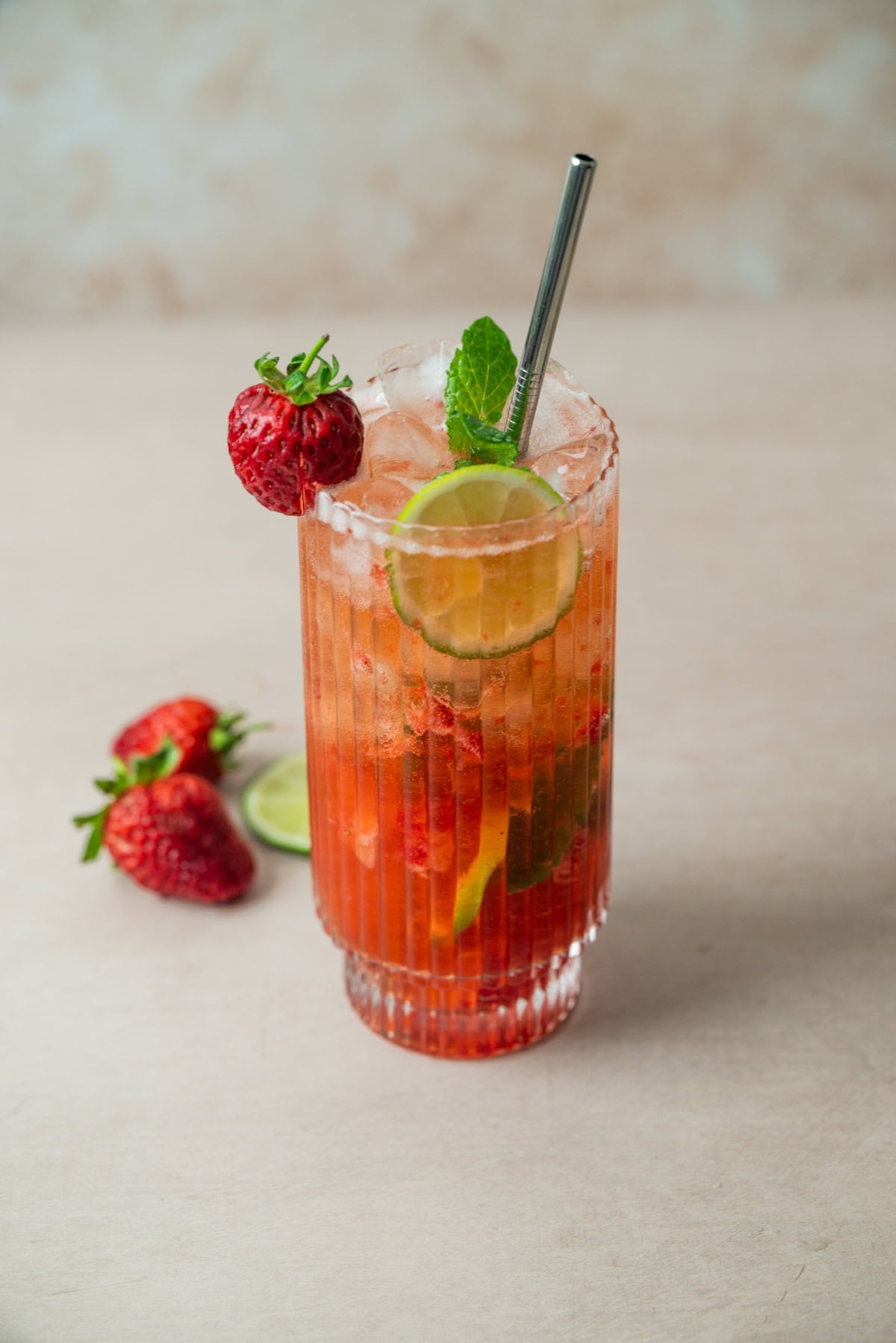 A tall glass of Strawberry Mojito with ice, garnished with a strawberry, lime slice, and mint sprig. A metal straw is in the glass. Two strawberries and a lime slice rest on the light surface nearby.