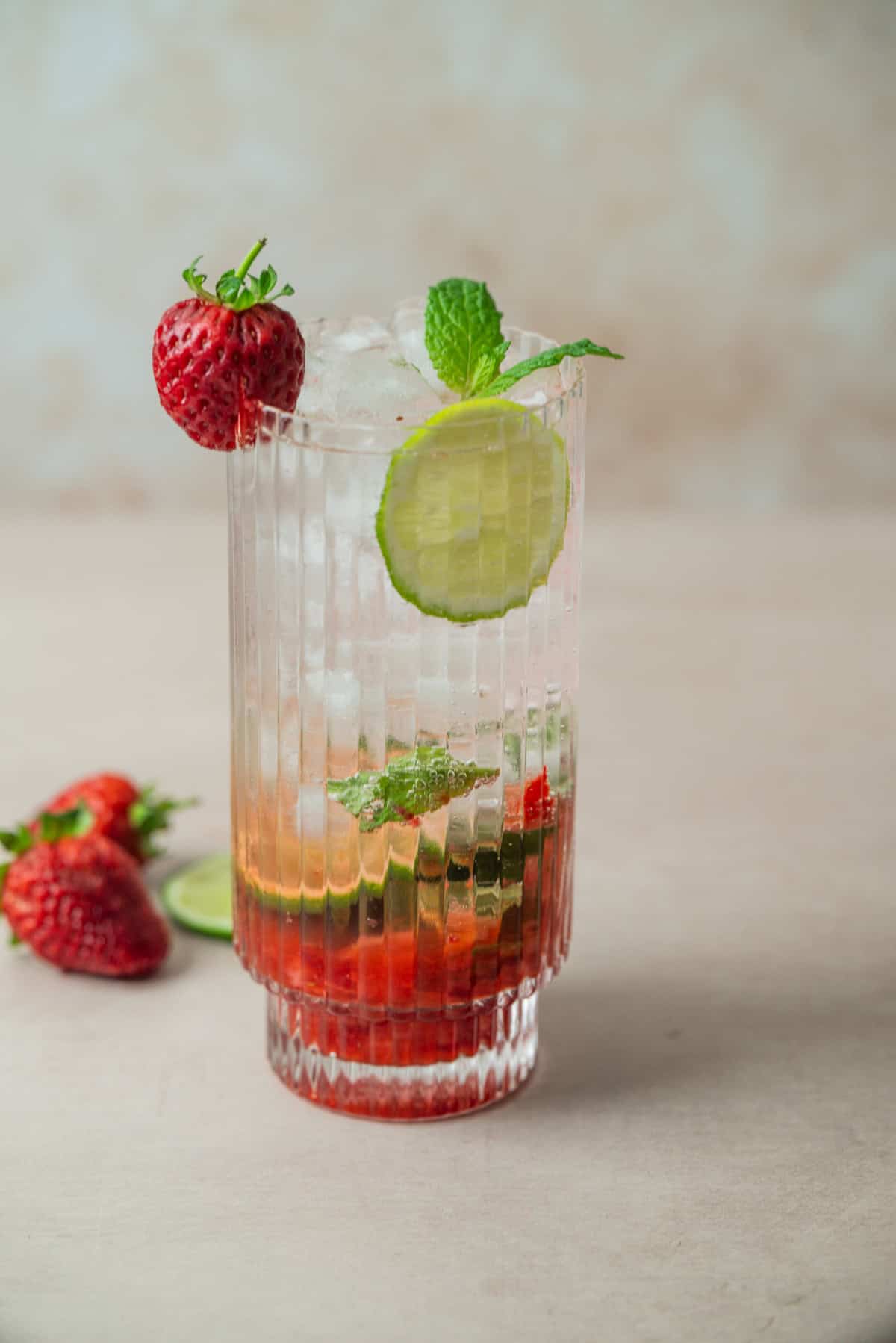 A tall, clear glass filled with a vibrant Strawberry Mojito garnished with a fresh strawberry, a lime slice, and a mint sprig. Ice cubes chill the drink, with strawberries and a lime slice beside the glass.