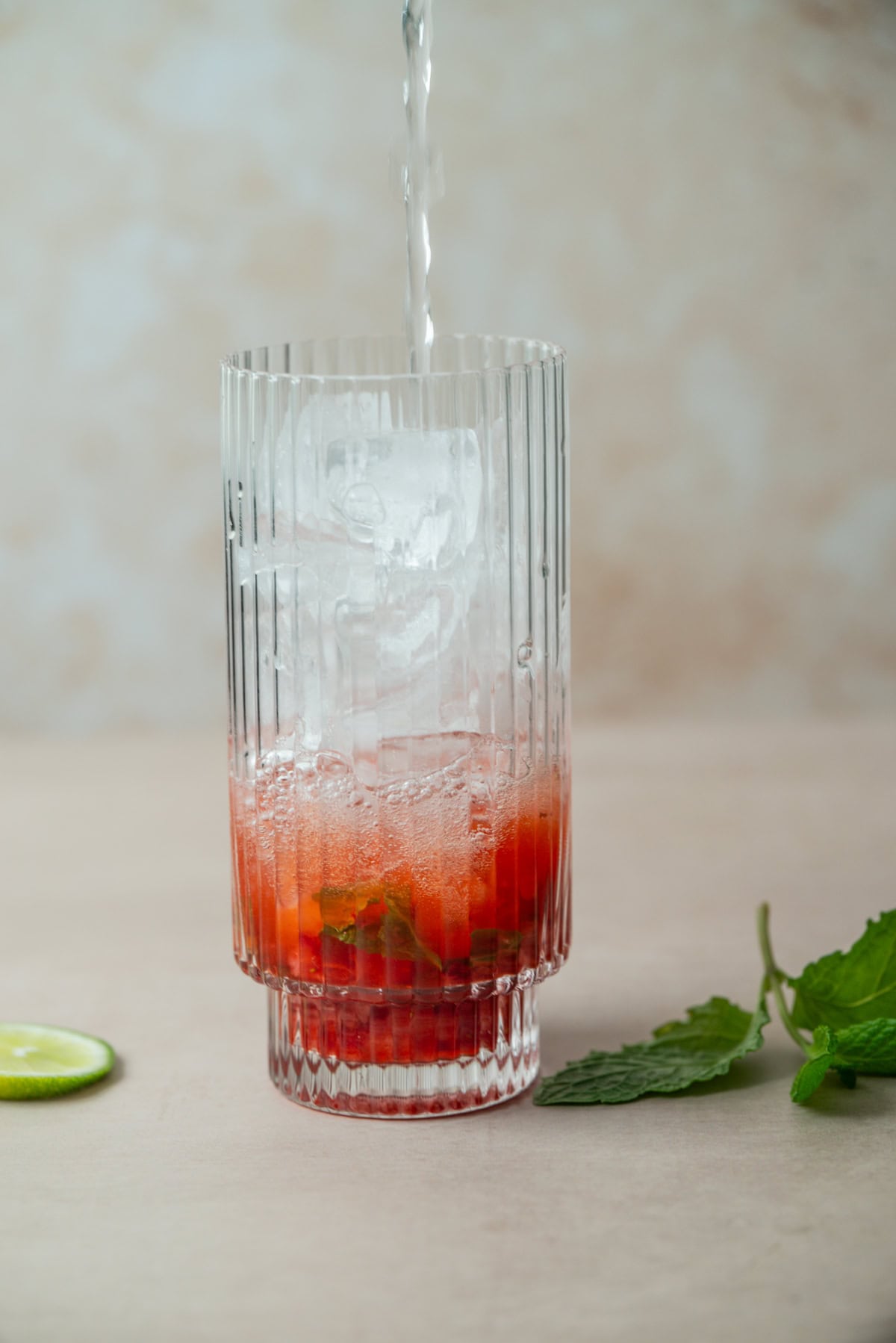 A clear, textured glass with ice, red fruit, and mint leaves is being filled with a clear liquid for a refreshing Strawberry Mojito. A lime slice and sprig of mint rest beside the glass on a beige surface.