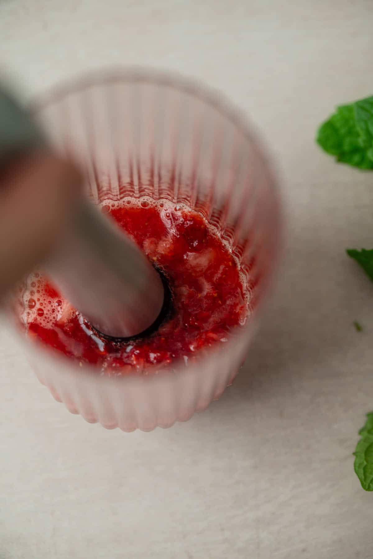 A close-up of a glass with red fruit being blended by an immersion blender, capturing the vibrant essence of a Strawberry Mojito. Fresh mint leaves are visible on the light-colored surface nearby.