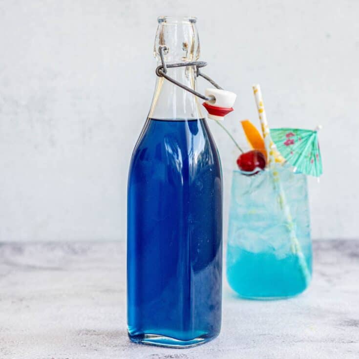 a blue lagoon mocktail in a tall glass with cherry, orange slice on rim and a straw, with a bottle of blue Curaçao syrup in front.