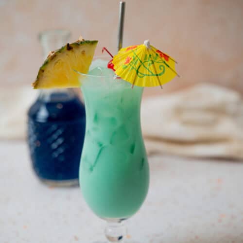 A tall glass of Blue Hawaiian Mocktail with ice, garnished with a pineapple slice, maraschino cherry, and a yellow paper umbrella, sits on a light countertop. A blue glass bottle is in the background.