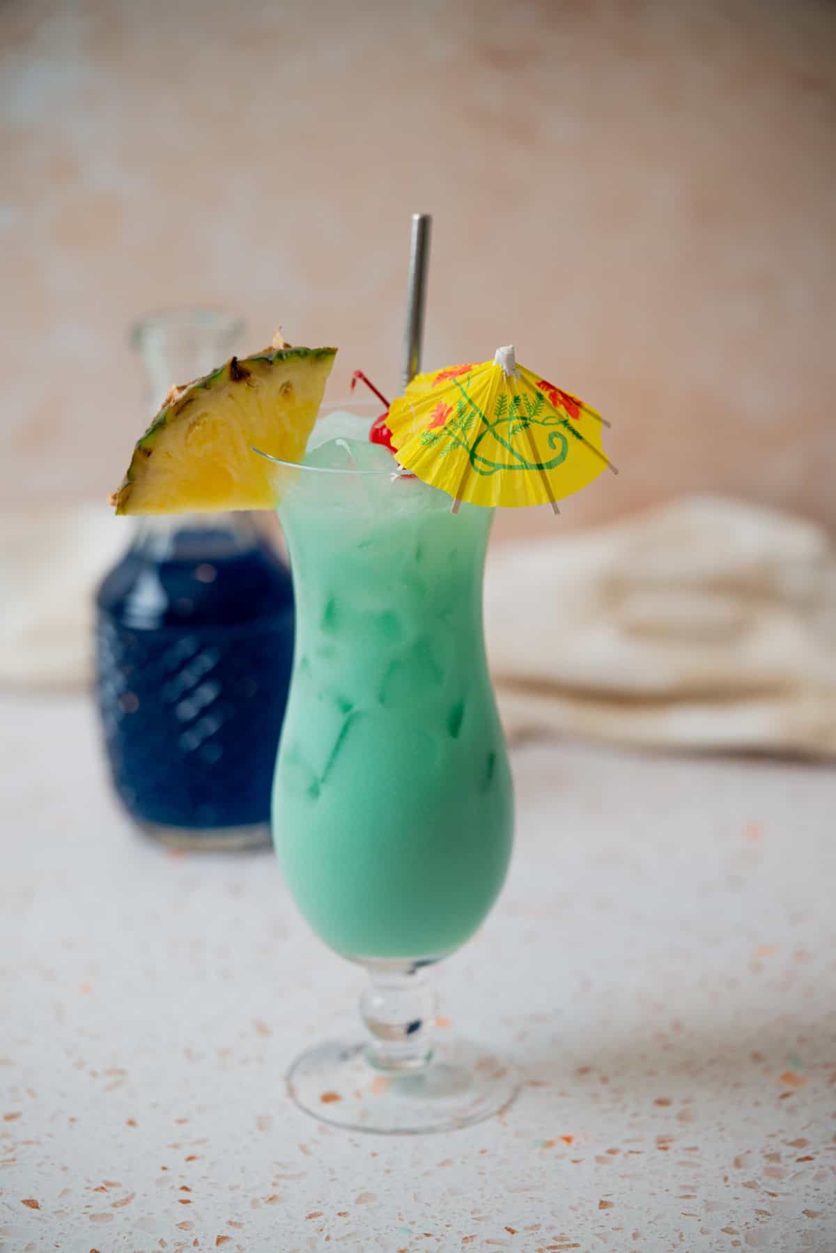 A tall glass of Blue Hawaiian Mocktail with ice, garnished with a pineapple slice, maraschino cherry, and a yellow paper umbrella, sits on a light countertop. A blue glass bottle is in the background.