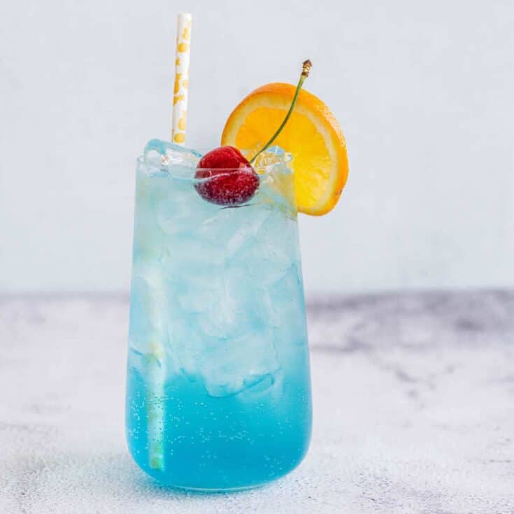 a blue lagoon mocktail in a tall glass with cherry, orange slice on rim and a straw, with a bottle of blue Curaçao syrup behind.