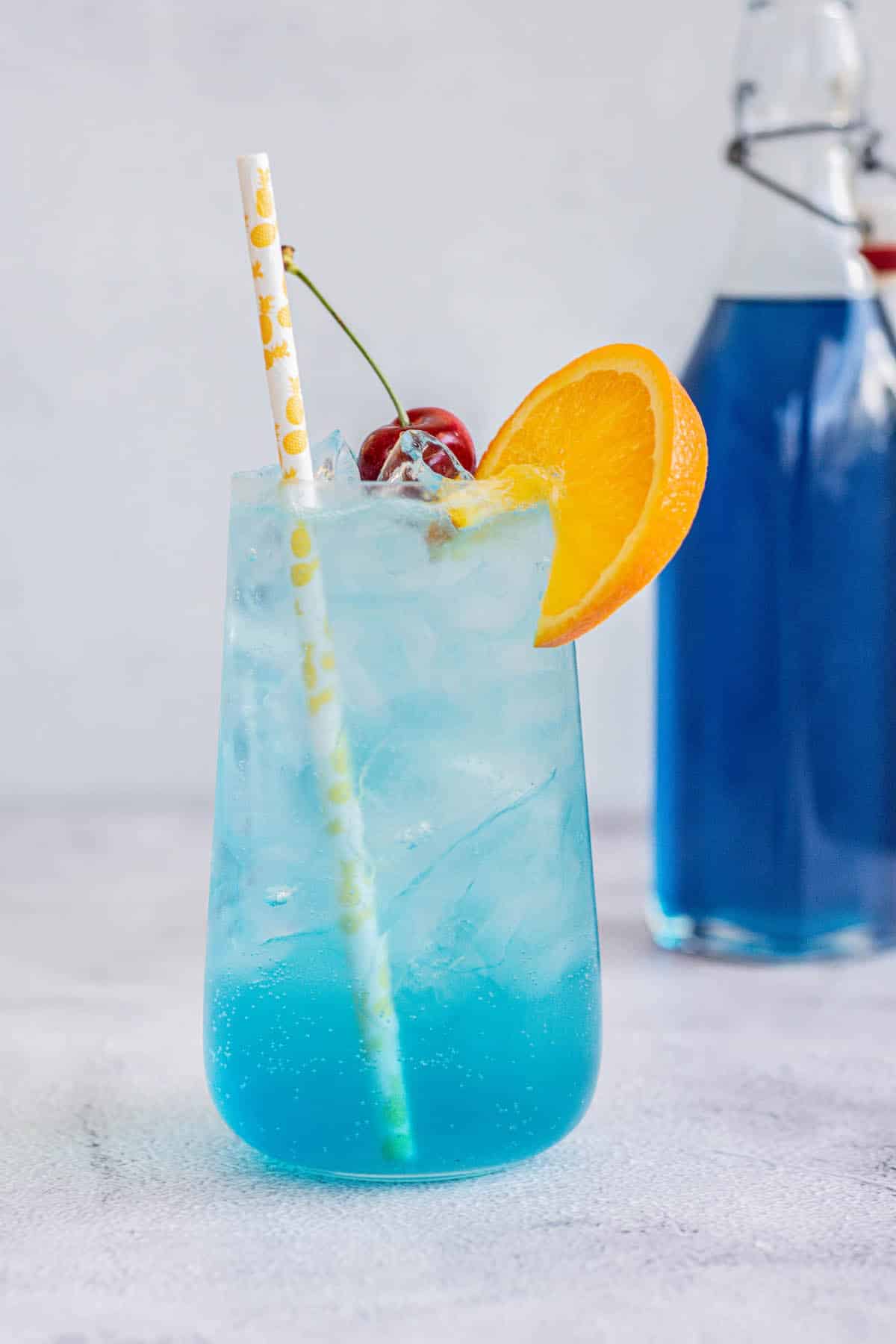 a blue lagoon mocktail in a tall glass with cherry, ice, orange slice on rim and a straw, with a bottle of blue Curaçao syrup behind.