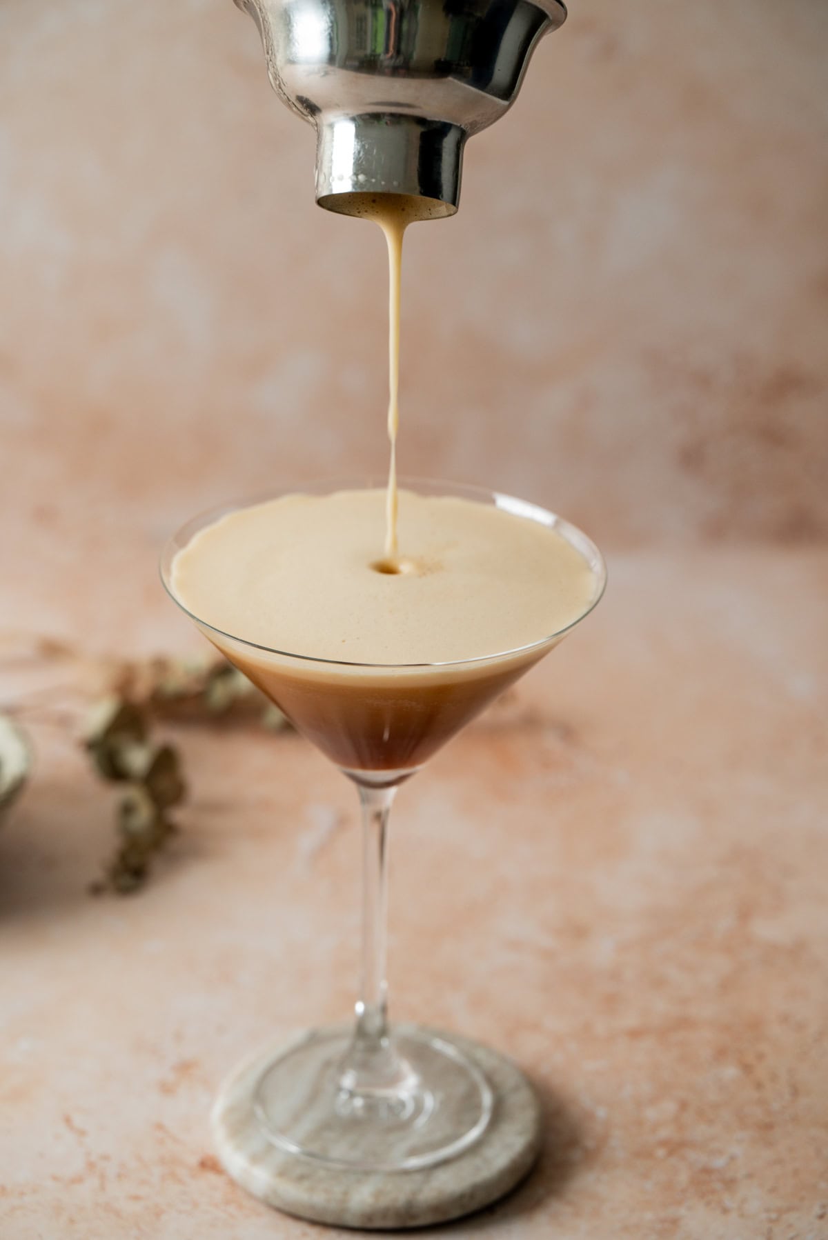 An Amaretto Espresso Martini is being poured from a shaker into a martini glass, creating a frothy layer on top. The glass sits on a marble coaster against a beige, softly blurred background.