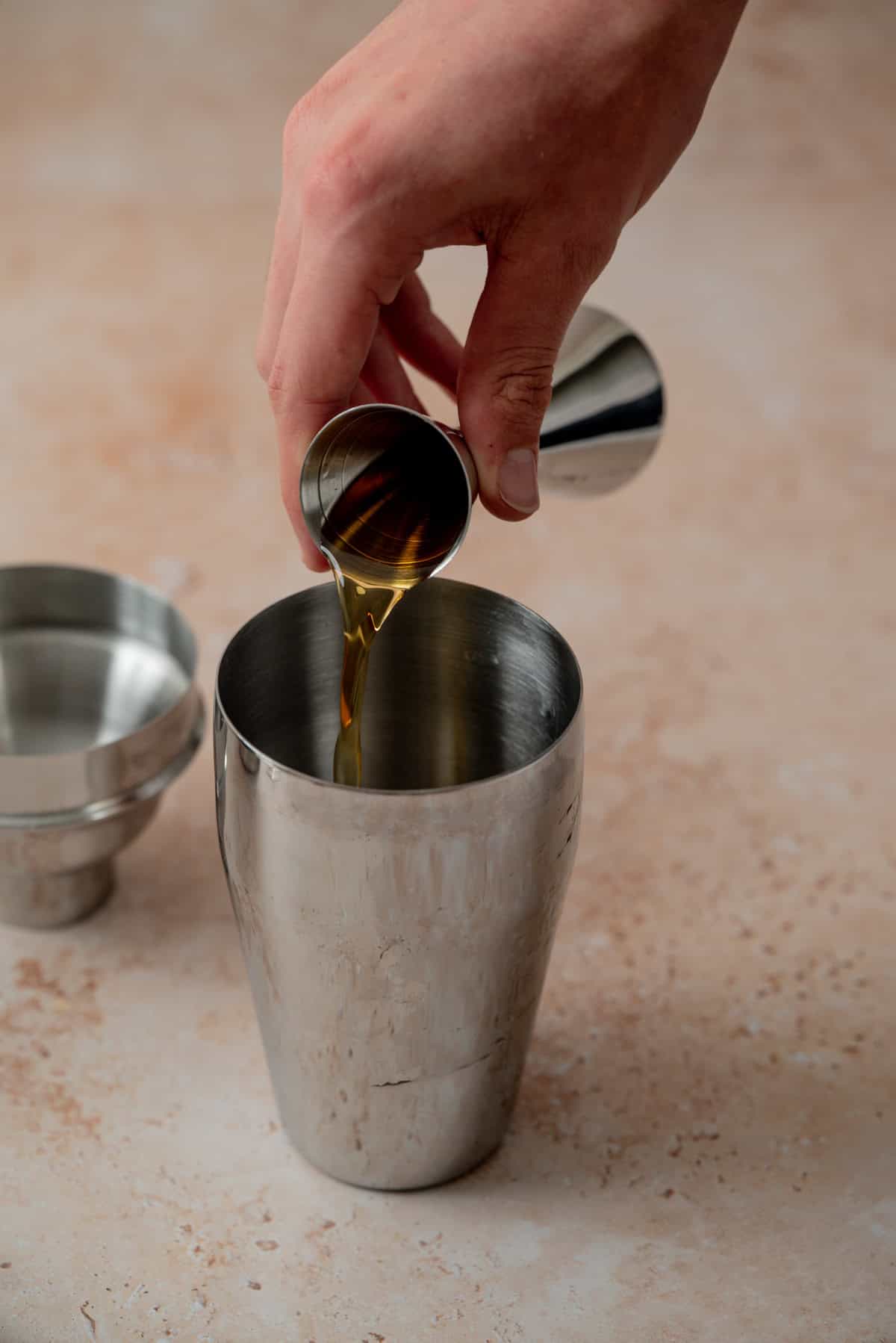 A hand pours a brown liquid from a small metal jigger into a stainless steel cocktail shaker on a light textured surface, preparing an Amaretto Espresso Martini. An empty jigger and cocktail shaker lid are nearby.