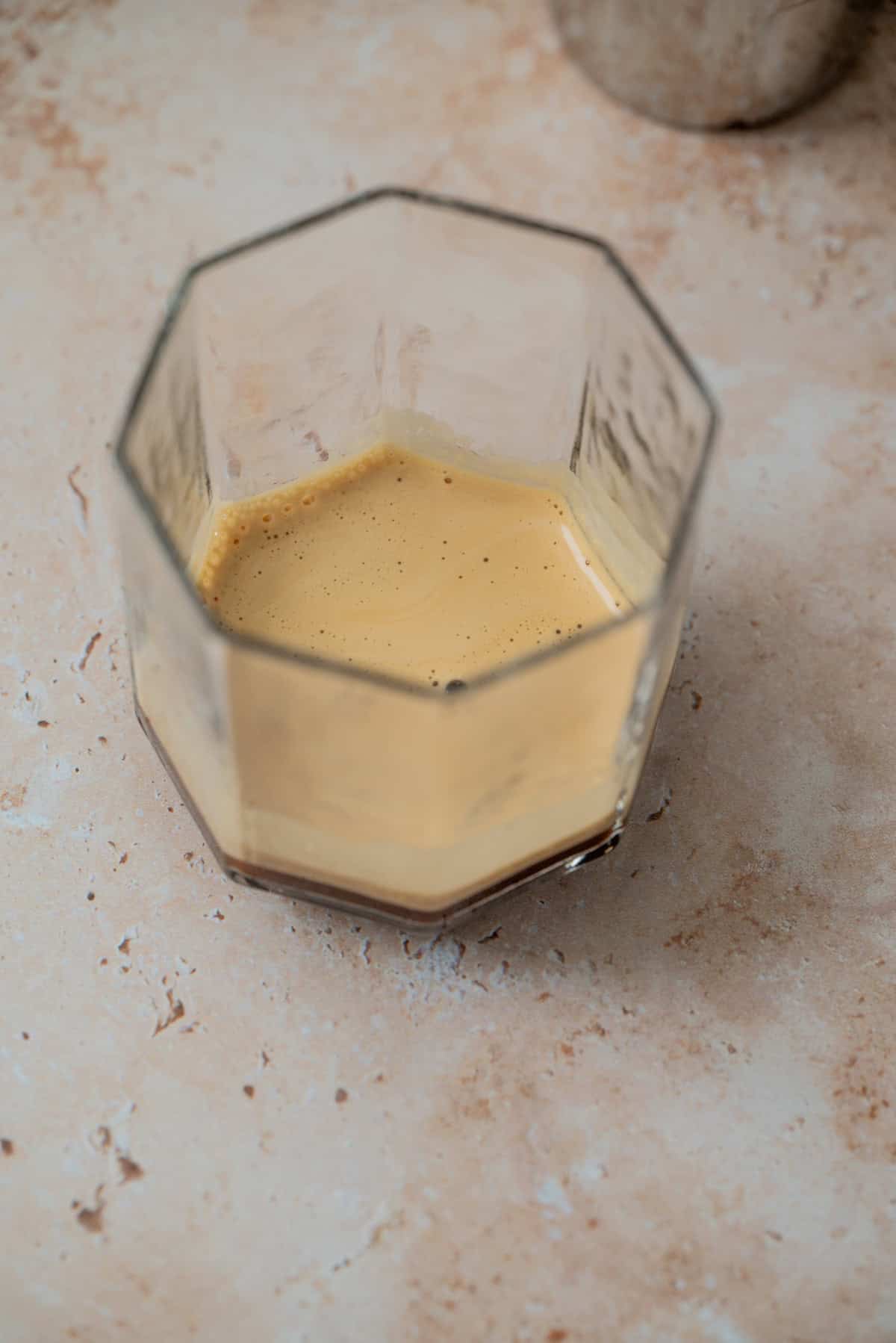 A glass with an octagonal shape containing a small amount of light brown Amaretto Espresso Martini or espresso crema sits on a beige textured surface.