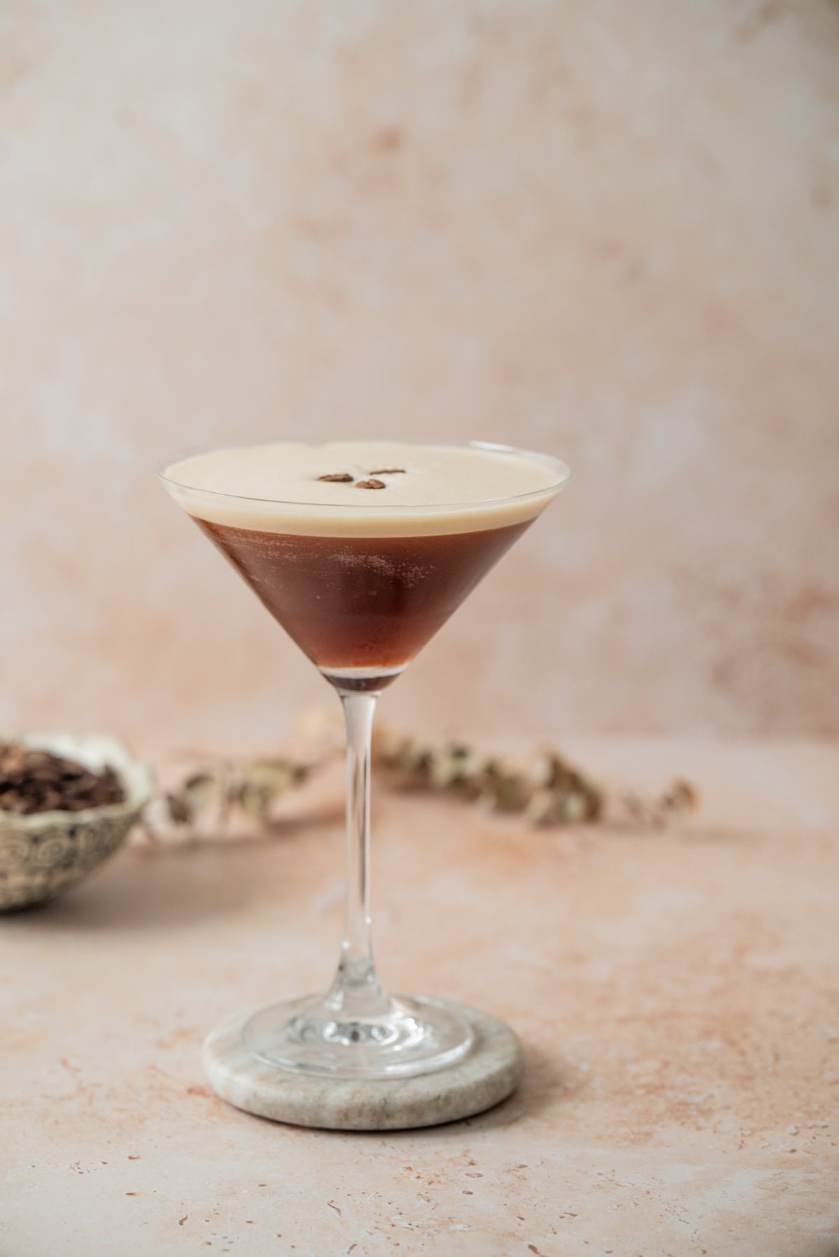 A martini glass filled with a dark Amaretto Espresso Martini, topped with creamy foam and three coffee beans, sits on a marble coaster against a light, textured background. A small bowl and dried flowers are blurred in the distance.