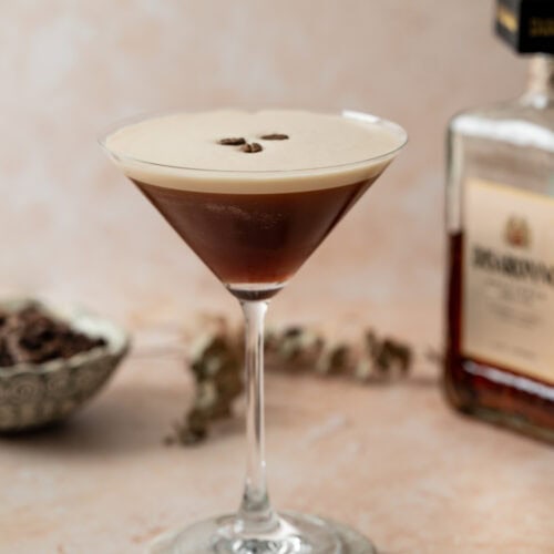 A martini glass filled with a dark Amaretto Espresso Martini, garnished with three coffee beans on top. A bottle and bowl are blurred in the background on a light, textured surface.