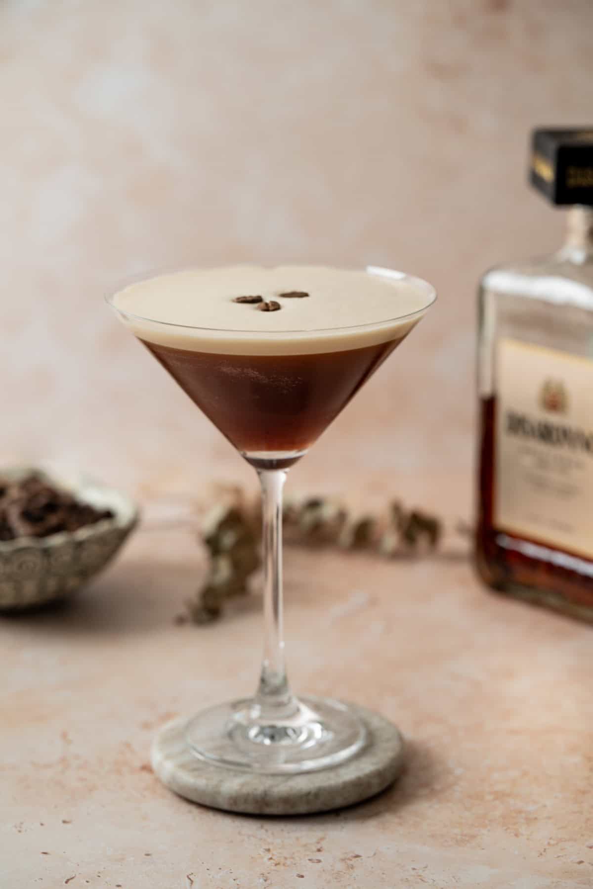 A martini glass filled with a dark Amaretto Espresso Martini, garnished with three coffee beans on top. A bottle and bowl are blurred in the background on a light, textured surface.